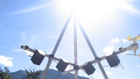 Photo of a metal triangular framework from which speakers and weather equipment is hung. The sun shines at the apex against a blue sky, with mountains in the background