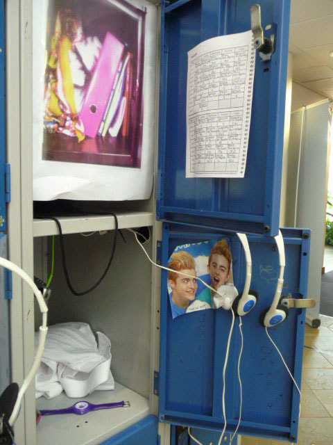 Blue metal school locker with three columns and four rows of lockers. Some doors are open with headphones hooked open them. The locker is decorated with images and pictures of the school and students interests.
