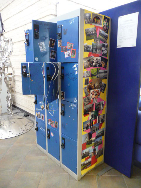 Blue metal school locker with three columns and four rows of lockers. Some doors are open with headphones hooked open them. The locker is decorated with images and pictures of the school and students interests.