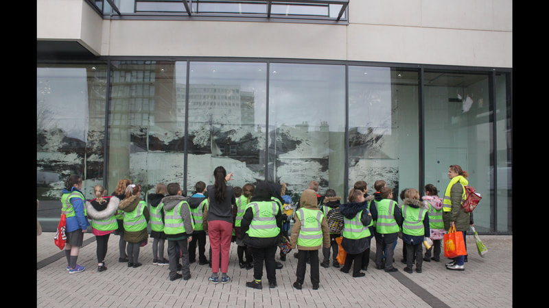 A group of children standing outside the gallery window, facing away from us. They are wearing green hi-viz jackets.
