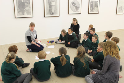 Damien sitting on gallery floor talking to pupils , who are sitting around her in a circle.