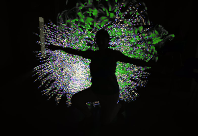 Silhouette of a woman facing to the left of the image with outstretched arms, and right leg raised. She is silhouetted by lines and sweeps of dotted light, mainly green.