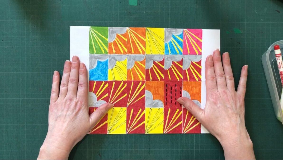 An overhead shot showing a white woman's hands resting on paper divided into coloured squares with repeating shapes. The background is a green cutting mat.Picture